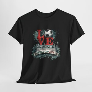May include: A black t-shirt featuring a graphic design with the word LOVE in red, where the O is replaced by a soccer ball. Below it, the text reads PHILADELPHIA and WORLD CUP 2026 on a banner, set against a blue-toned city skyline silhouette.