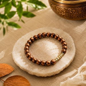 Peut inclure: Un bracelet circulaire composé de perles sphériques polies de couleur cuivre métallique. Le bracelet présente une perle d'accent cylindrique unique avec une texture dorée et nervurée, positionnée le long du rang de perles rondes.