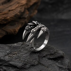 Könnte beinhalten: Ein silberfarbener Metallring mit einem detaillierten Design aus drei scharfen, texturierten Krallen, die sich um den Finger winden. Die Oberfläche der Krallen zeigt ein schuppiges, reptilienartiges Muster mit polierten Spitzen und einer glatten Ringschiene.