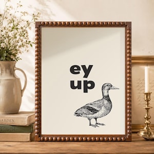 May include: A framed wall art print featuring a black and white line drawing of a duck positioned below the bold black text "ey up". The artwork is encased in a dark brown wooden frame with a decorative beaded texture along the outer edge.
