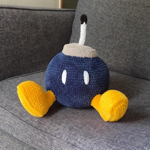 Puede incluir: Un peluche con forma de bomba redonda con un cuerpo texturizado azul oscuro, dos pies de color amarillo brillante y una sección superior beige. Una mecha blanca y negra sobresale del centro. La bomba tiene dos ojos ovalados blancos al frente.