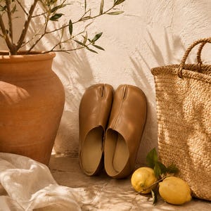 May include: A pair of tan leather slip-on shoes featuring a distinct split-toe design. The shoes have a smooth, matte finish with a low-profile silhouette, a rounded toe box, and a central seam running from the split to the opening of the shoe.