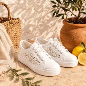 Può includere: Un paio di sneakers basse bianche con un motivo floreale tagliato al laser sui pannelli laterali, lacci bianchi e una spessa suola in gomma. Le scarpe presentano una finitura liscia e occhielli color argento per la chiusura con lacci.