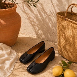 May include: A pair of black leather ballet flats featuring a distinct split-toe design at the front. These shoes have a low block heel, a smooth finish, and a simple slip-on construction with a small central notch detail on the vamp.