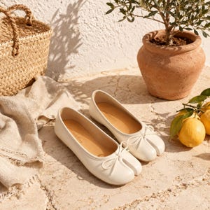 Könnte beinhalten: Ein Paar cremeweiße Ballerinas mit geteilter Zehenpartie, glatter lederartiger Oberfläche, abgerundeter Öffnung und einer zarten Schleife an der Vorderseite. Das Innere der Schuhe ist mit beigem Material ausgekleidet und trägt einen Markennamen.