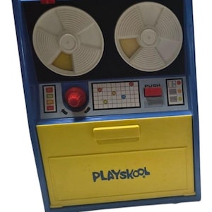 May include: A vintage Playskool toy computer with a blue frame and a large yellow drawer. The top section features two white circular reels, a red dial, a grid panel with colored dots, a red push button, and a small screen with colored squares.