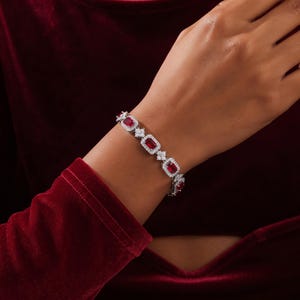 May include: A silver-toned link bracelet featuring rectangular red gemstones, each surrounded by a halo of small clear stones. The links are connected by small, four-petaled floral-shaped accents set with clear stones, creating a continuous pattern.