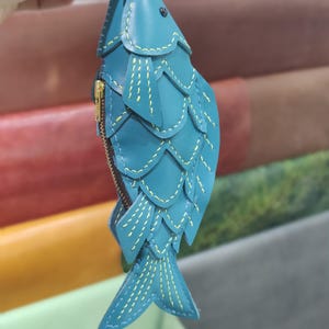 Op de afbeelding: Een blauwgroene, visvormige portemonnee van leer met gelaagde, schubachtige panelen die zijn gestikt met geel garen. Het accessoire is voorzien van een goudkleurige metalen rits aan de zijkant en een klein donker oogdetail aan de bovenzijde.