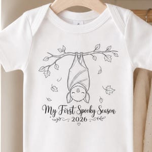 May include: A white short-sleeved baby bodysuit featuring a black line art illustration of a bat hanging from a branch surrounded by leaves. Below the bat, the text reads "My First Spooky Season" with the year "2026" printed underneath.