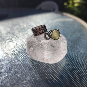 May include: Two silver-toned rings resting on a clear, textured block. One ring features a wide band with an intricate, repeating geometric pattern, while the other is a thinner, wire-wrapped band holding a small, pale yellow, translucent stone.