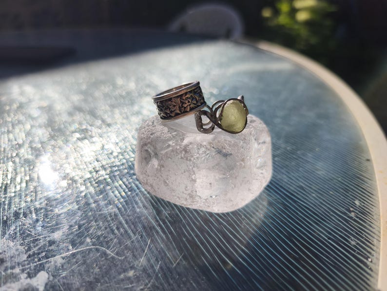 May include: Two silver-toned rings resting on a clear, textured block. One ring features a wide band with an intricate, repeating geometric pattern, while the other is a thinner, wire-wrapped band holding a small, pale yellow, translucent stone.