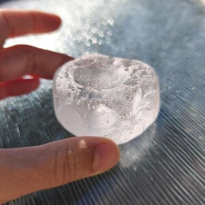 May include: A clear, circular glass object with a textured, uneven surface and a shallow, concave depression in the center. The piece has a translucent, crystalline appearance with visible internal fractures and a smooth, rounded outer edge.