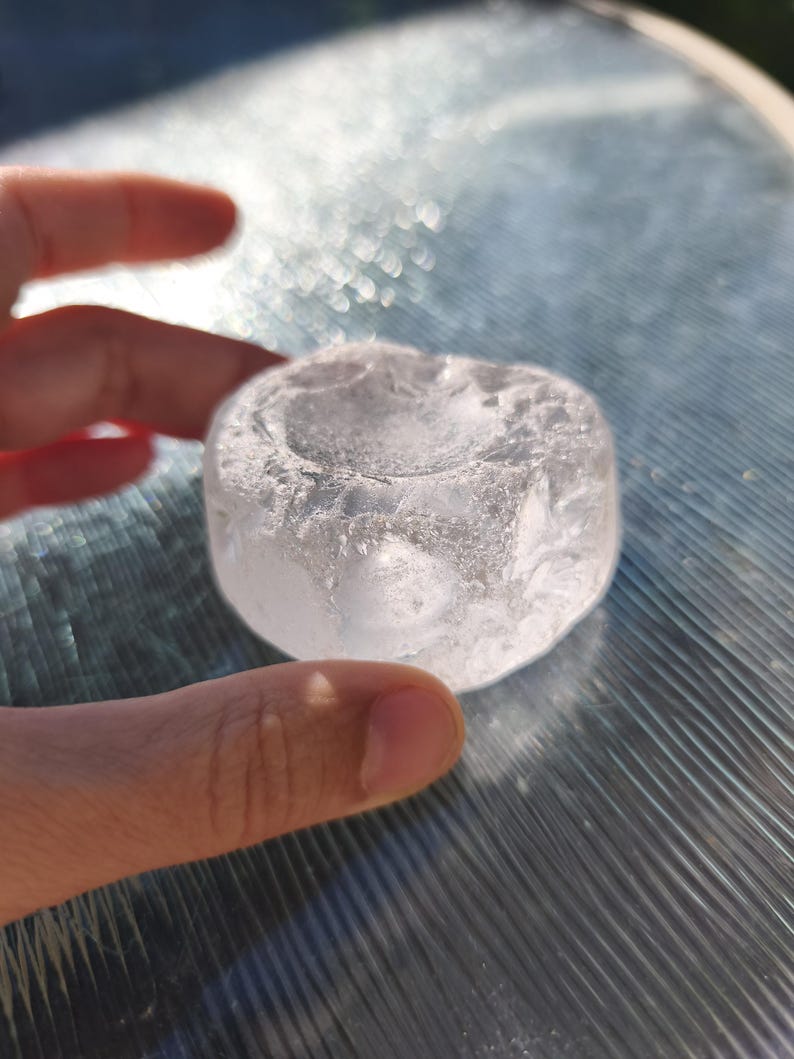 May include: A clear, circular glass object with a textured, uneven surface and a shallow, concave depression in the center. The piece has a translucent, crystalline appearance with visible internal fractures and a smooth, rounded outer edge.
