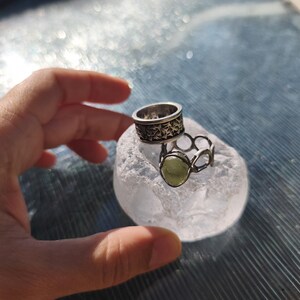 May include: Two silver-toned rings resting on a translucent, textured white stone. One ring features a wide band with a dark, intricate etched pattern, while the other is a thinner, open-link band set with a single, smooth, pale green oval stone.