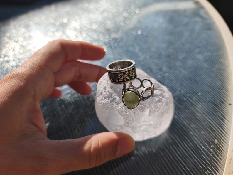 May include: Two silver-toned rings resting on a translucent, textured white stone. One ring features a wide band with a dark, intricate etched pattern, while the other is a thinner, open-link band set with a single, smooth, pale green oval stone.