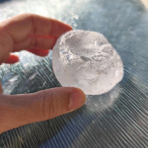 May include: A clear, translucent, irregularly shaped stone with a smooth, polished surface and a shallow, concave indentation on the top side. The object has a crystalline appearance with subtle internal textures and light-reflecting facets.
