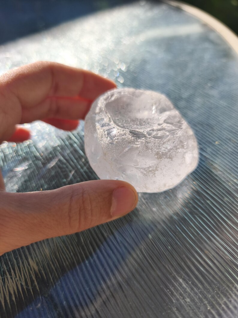 May include: A clear, translucent, irregularly shaped stone with a smooth, polished surface and a shallow, concave indentation on the top side. The object has a crystalline appearance with subtle internal textures and light-reflecting facets.
