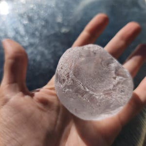 May include: A translucent, pale pink, rounded stone with a smooth, polished surface and irregular, textured edges. The object has a semi-transparent appearance, showing internal fractures and variations in clarity throughout its solid form.