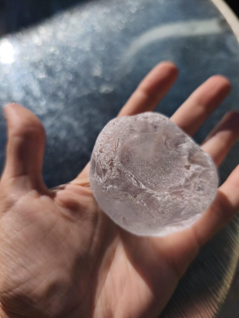 May include: A translucent, pale pink, rounded stone with a smooth, polished surface and irregular, textured edges. The object has a semi-transparent appearance, showing internal fractures and variations in clarity throughout its solid form.