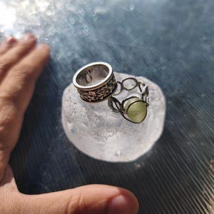 May include: Two silver-toned rings resting on a translucent, textured, circular base. One ring features a wide band with an intricate, repeating star-like pattern. The second ring is constructed from thin wire loops holding a pale green, round stone.