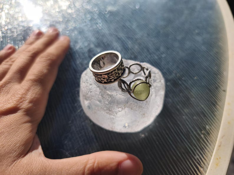 May include: Two silver-toned rings resting on a translucent, textured, circular base. One ring features a wide band with an intricate, repeating star-like pattern. The second ring is constructed from thin wire loops holding a pale green, round stone.