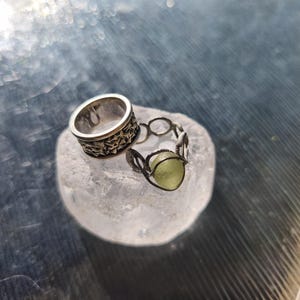 May include: Two silver rings resting on a translucent white stone. One ring is a wide band featuring a repeating star pattern. The second ring is a delicate wire-wrapped design holding a single, small, round, pale green stone in its center.