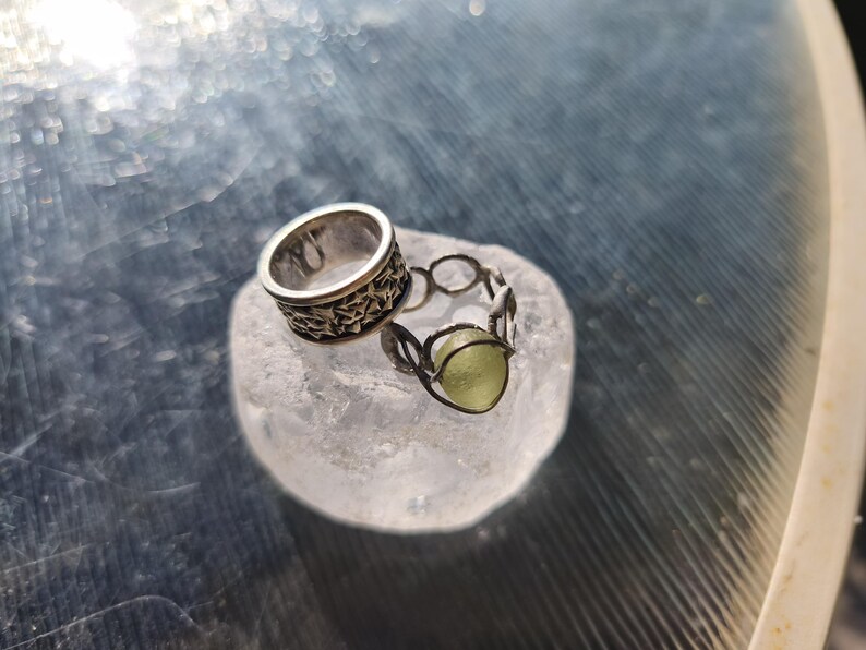 May include: Two silver rings resting on a translucent white stone. One ring is a wide band featuring a repeating star pattern. The second ring is a delicate wire-wrapped design holding a single, small, round, pale green stone in its center.