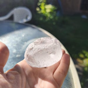May include: A clear, translucent, rounded glass object with a textured, uneven surface and a shallow circular indentation on the top. The piece has a smooth, polished appearance in some areas and a rough, frosted texture along the outer edges.