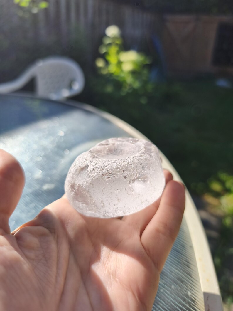 May include: A clear, translucent, rounded glass object with a textured, uneven surface and a shallow circular indentation on the top. The piece has a smooth, polished appearance in some areas and a rough, frosted texture along the outer edges.