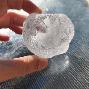 May include: A clear, irregularly shaped piece of sea glass with a smooth, frosted texture. The object is translucent, allowing light to pass through its rounded edges and uneven surface, displaying a cloudy, white interior with subtle variations.