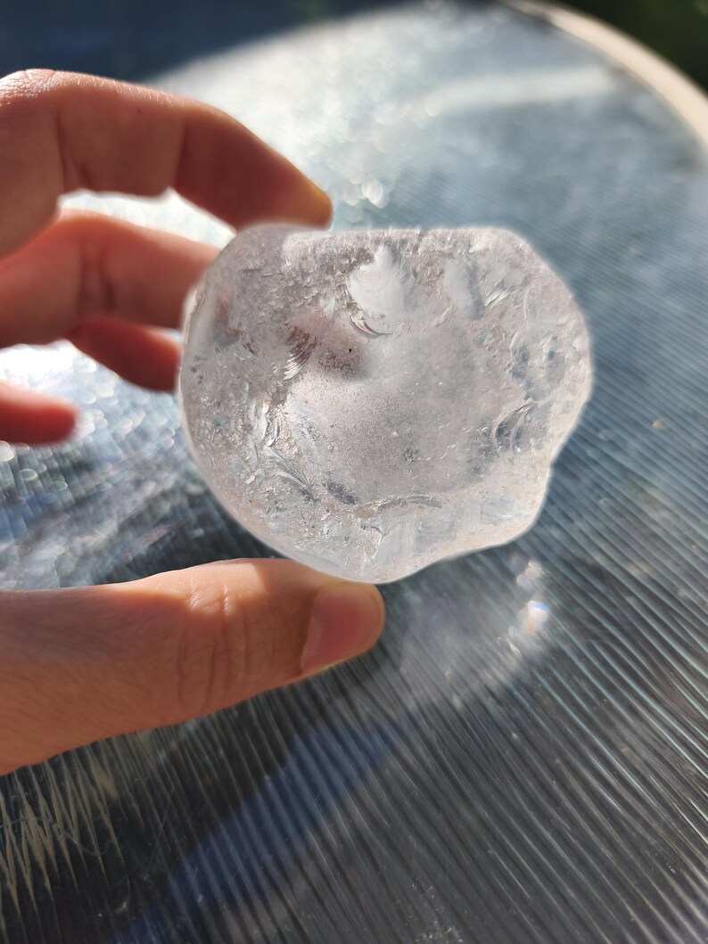 May include: A clear, irregularly shaped piece of sea glass with a smooth, frosted texture. The object is translucent, allowing light to pass through its rounded edges and uneven surface, displaying a cloudy, white interior with subtle variations.