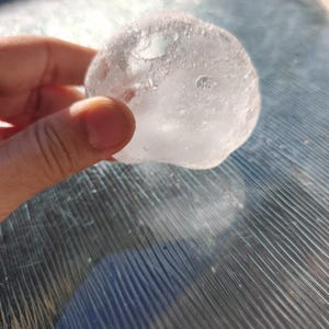 May include: A clear, rounded, and translucent object with a textured, uneven surface. The material appears glass-like and solid, catching the sunlight to create bright highlights and internal reflections throughout its irregular, organic shape.
