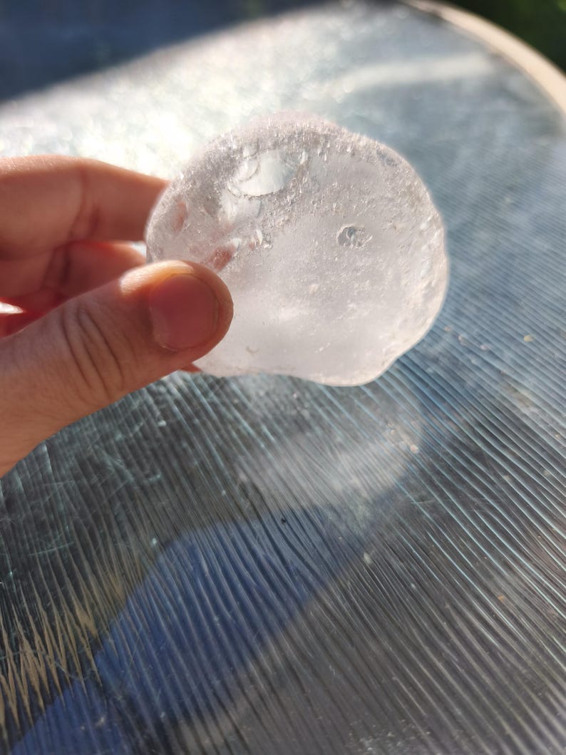 May include: A clear, rounded, and translucent object with a textured, uneven surface. The material appears glass-like and solid, catching the sunlight to create bright highlights and internal reflections throughout its irregular, organic shape.