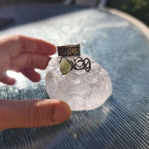 May include: Two silver rings stacked on a clear, irregular crystal. The top ring features a dark, etched floral pattern, while the bottom ring has an open, organic wire design holding a small, pale green, teardrop-shaped stone.