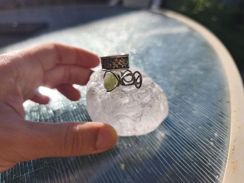 May include: Two silver rings stacked on a clear, irregular crystal. The top ring features a dark, etched floral pattern, while the bottom ring has an open, organic wire design holding a small, pale green, teardrop-shaped stone.