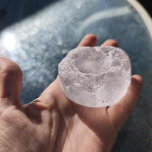 May include: A circular, translucent, and colorless object with a rough, uneven texture and a shallow, concave depression in the center. The item appears to be made of a solid, clear material with a slightly frosted or crystalline surface finish.