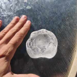 May include: A clear, irregularly shaped piece of raw quartz crystal with a translucent, glass-like appearance and a textured, multifaceted surface. The stone has a rounded, organic form with visible internal fractures and light-reflecting edges.
