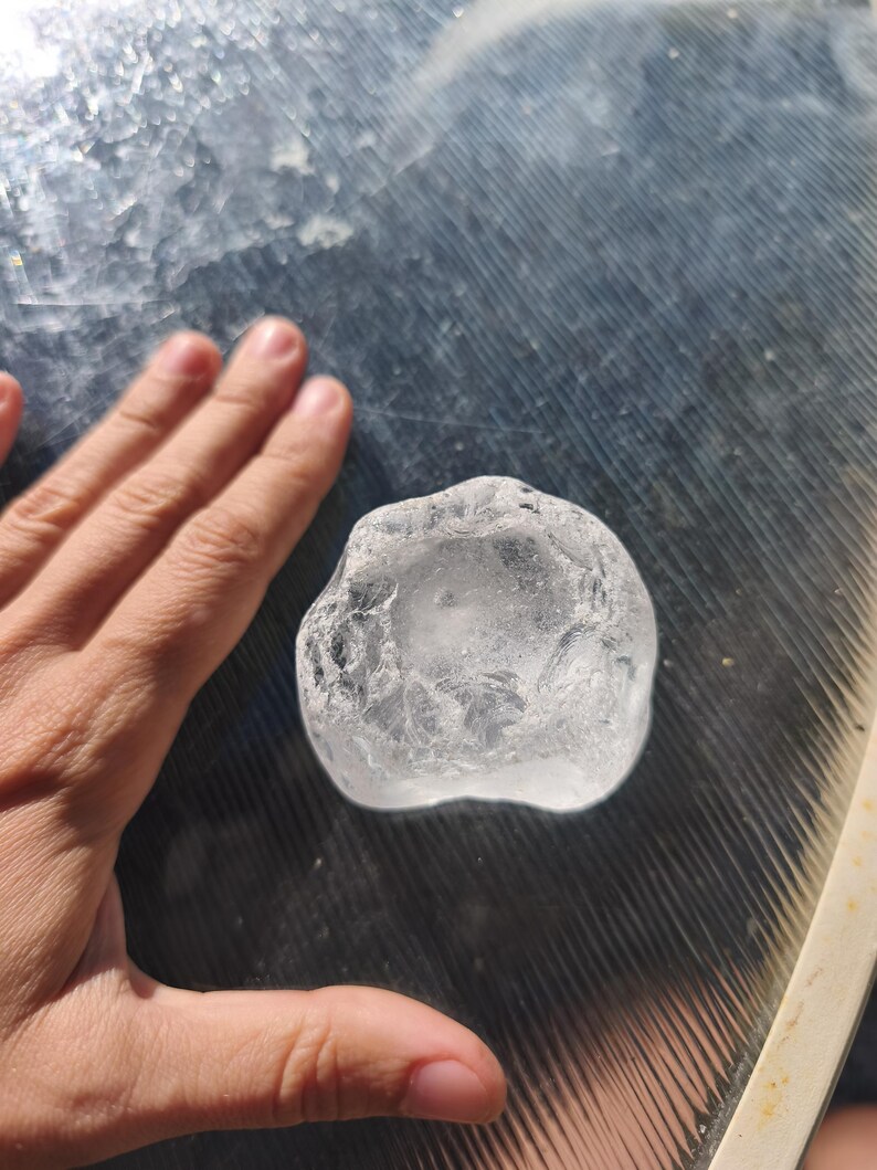 May include: A clear, irregularly shaped piece of raw quartz crystal with a translucent, glass-like appearance and a textured, multifaceted surface. The stone has a rounded, organic form with visible internal fractures and light-reflecting edges.