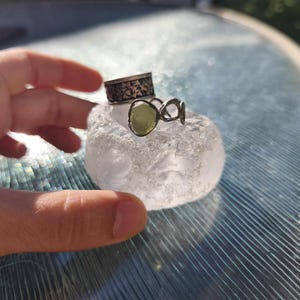 May include: A silver ring featuring a textured band and a second ring with an open, organic metal design holding a single, pale green, translucent stone. Both rings are resting on a clear, textured, rounded piece of glass or crystal.