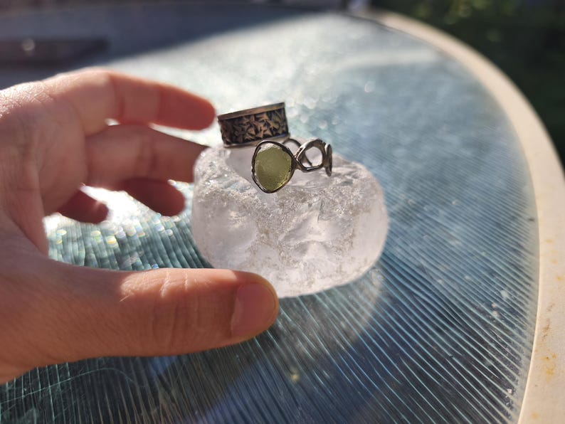 May include: A silver ring featuring a textured band and a second ring with an open, organic metal design holding a single, pale green, translucent stone. Both rings are resting on a clear, textured, rounded piece of glass or crystal.