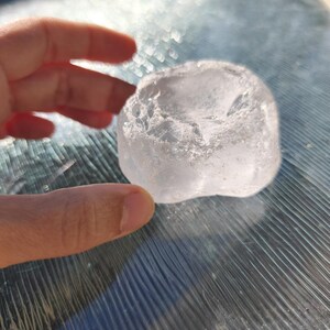 May include: A clear, translucent, irregularly shaped piece of raw quartz crystal with a rough, textured surface and a slightly concave top. The stone exhibits natural facets and internal inclusions that catch the light, creating a bright appearance.