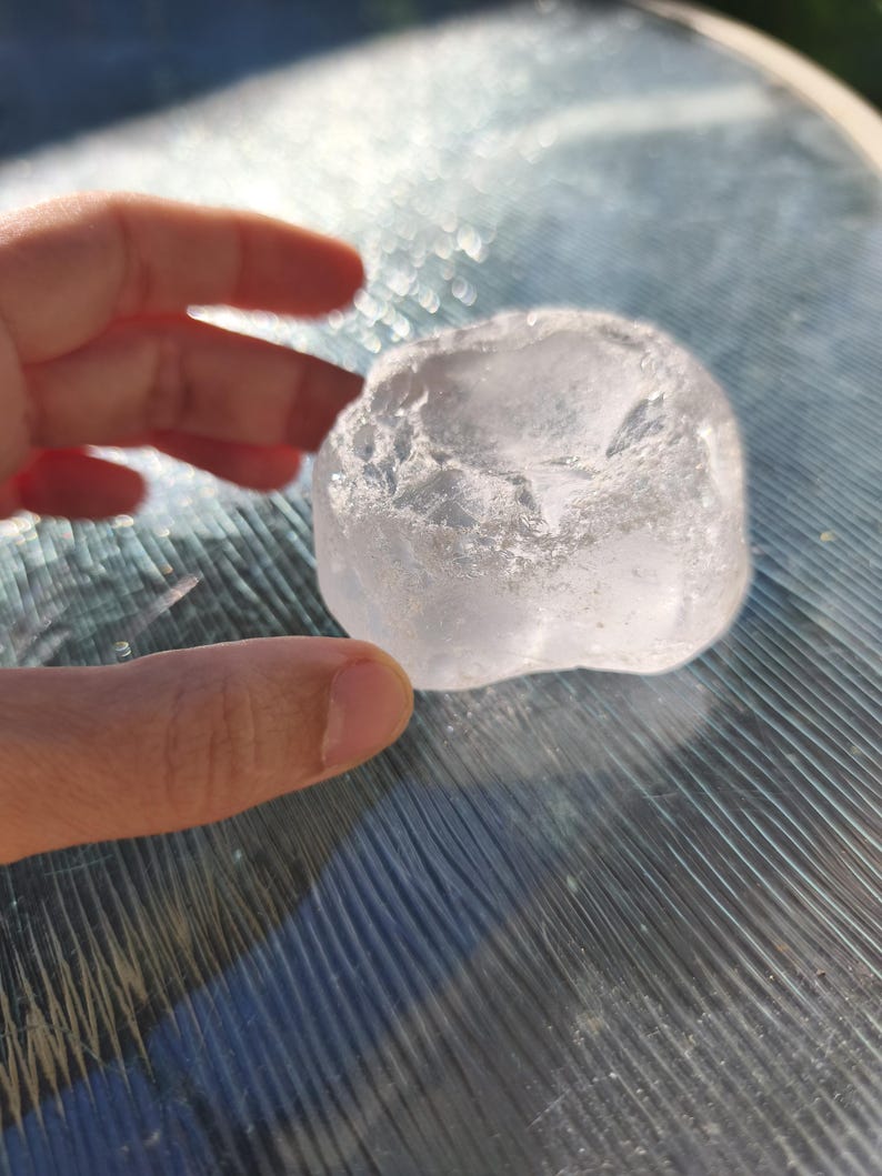May include: A clear, translucent, irregularly shaped piece of raw quartz crystal with a rough, textured surface and a slightly concave top. The stone exhibits natural facets and internal inclusions that catch the light, creating a bright appearance.