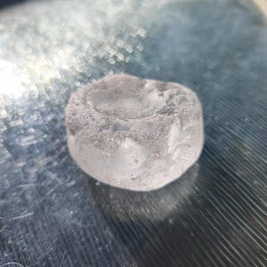May include: A small, translucent, and colorless circular object with a smooth, slightly concave top surface and a textured, uneven outer edge. The material appears clear like glass or ice, featuring internal inclusions and a polished, rounded form.