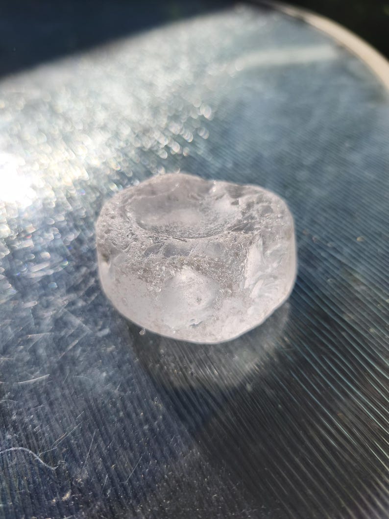 May include: A small, translucent, and colorless circular object with a smooth, slightly concave top surface and a textured, uneven outer edge. The material appears clear like glass or ice, featuring internal inclusions and a polished, rounded form.