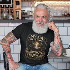Könnte beinhalten: Ein schwarzes Kurzarm-T-Shirt mit einem Grafikdruck eines goldenen Audio-Lautstärkereglers und dem Text MY AGE MY BUSINESS MY VOLUME EVERYBODY'S BUSINESS. Das Design enthält verzierte Schnörkel um die zentrale Regler-Grafik.