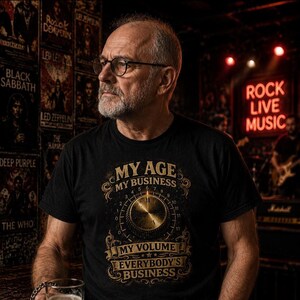 Może przedstawiać: Czarny t-shirt ze złotą grafiką przedstawiającą okrągłe pokrętło głośności. Tekst brzmi: MY AGE MY BUSINESS MY VOLUME EVERYBODY'S BUSINESS. Wzór zawiera ozdobne zawijasy otaczające centralną grafikę pokrętła.