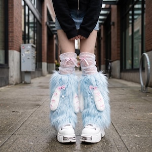May include: Light blue faux fur leg warmers featuring white plush bunny ears with pink bows, worn over white polka-dot sheer ruffled socks. The leg warmers are paired with white platform shoes that have metallic silver hardware details on the front.