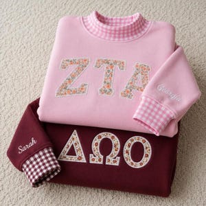 Peut inclure: Un sweat-shirt rose clair avec une applique florale ZTA et un col vichy, et un sweat-shirt bordeaux avec une applique florale Delta Omega. La manche rose porte le nom Georgia, et la manche bordeaux porte le nom Sarah.
