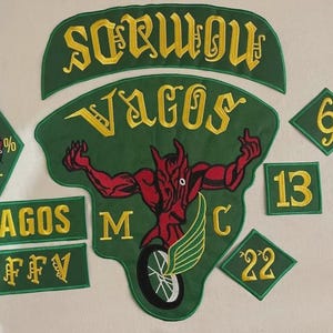 May include: A collection of green and yellow embroidered patches featuring the text "VAGOS", "NOMADS", "VFFV", "MC", "1%", "6", "13", and "22". The central patch displays a red devil figure riding a motorcycle wheel with wings, surrounded by yellow lettering.