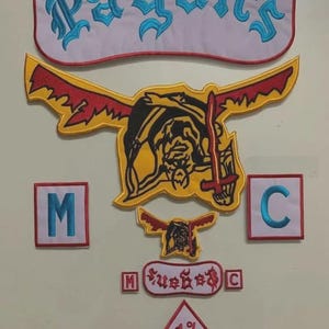 May include: A collection of embroidered patches including a large white rocker patch with the blue text Pagan's, a large yellow and red winged figure patch, two square patches with blue letters M and C, a smaller winged figure, and a diamond 1% patch.